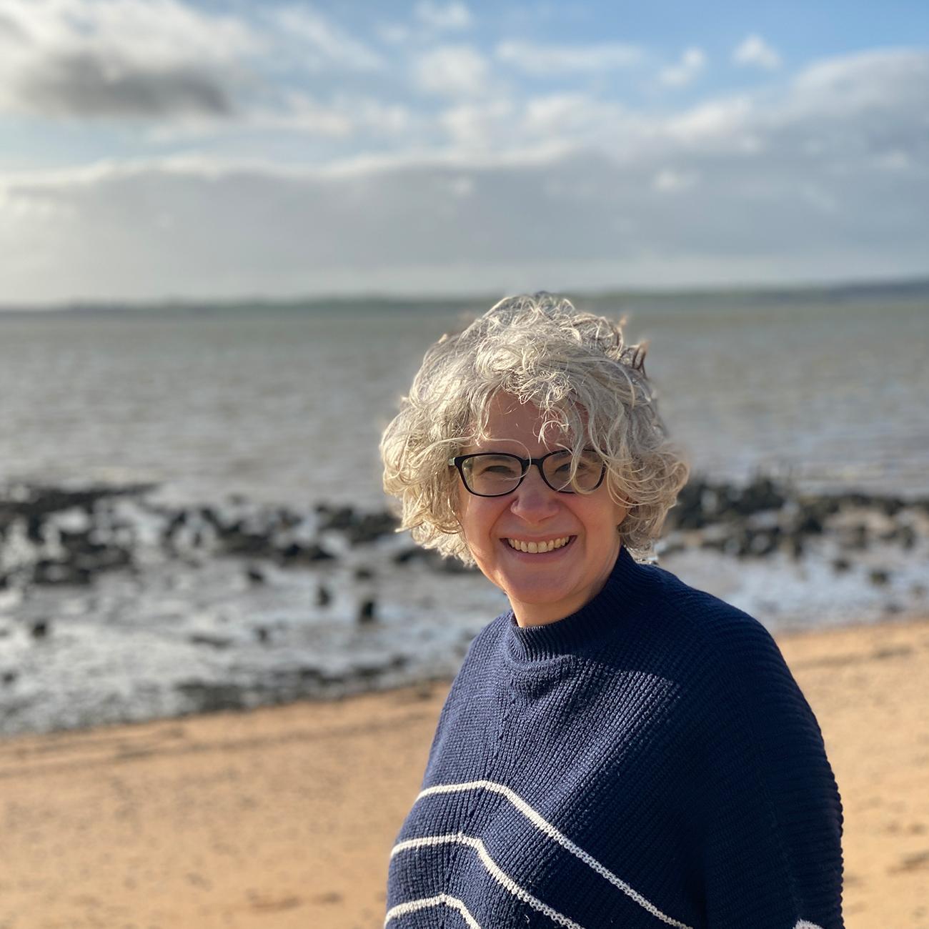 picture of a woman on the beach wearing glasses and a navy and white striped jumper.  She has short, grey hair